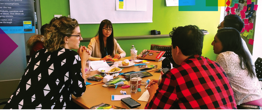 A group of young adults sits around a table in a discussion, with papers, booklets, and snacks spread out in front of them. The participants appear engaged in conversation, and one person listens attentively while others speak. The background features a green wall, sticky notes, and a poster with motivational text. The setting suggests a collaborative workshop focused on youth-adult engagement.
